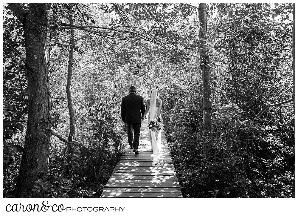 black and white photo of a bride and groom walking down a boardwalk in the woods