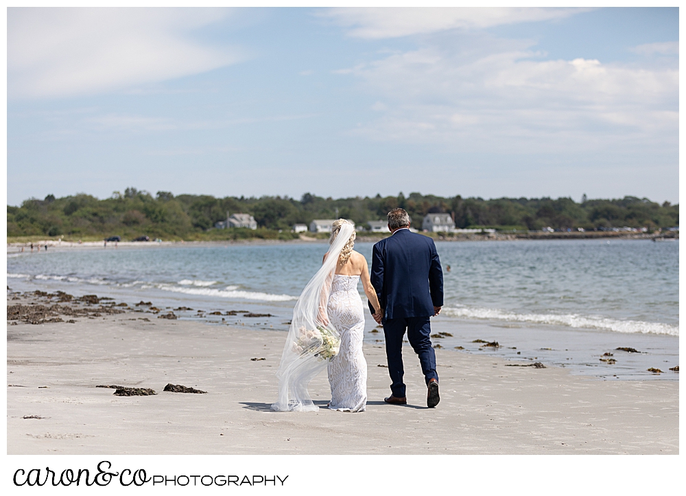 bride and groom, their backs to the camera, walk along the beach at the water's edge