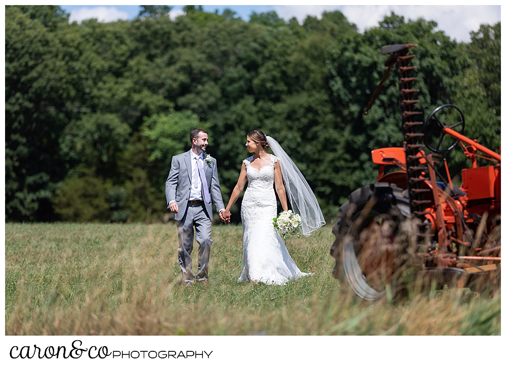 sweet summertime wedding photo of a bride and groom walking hand in hand in a field, next to an orange tractor