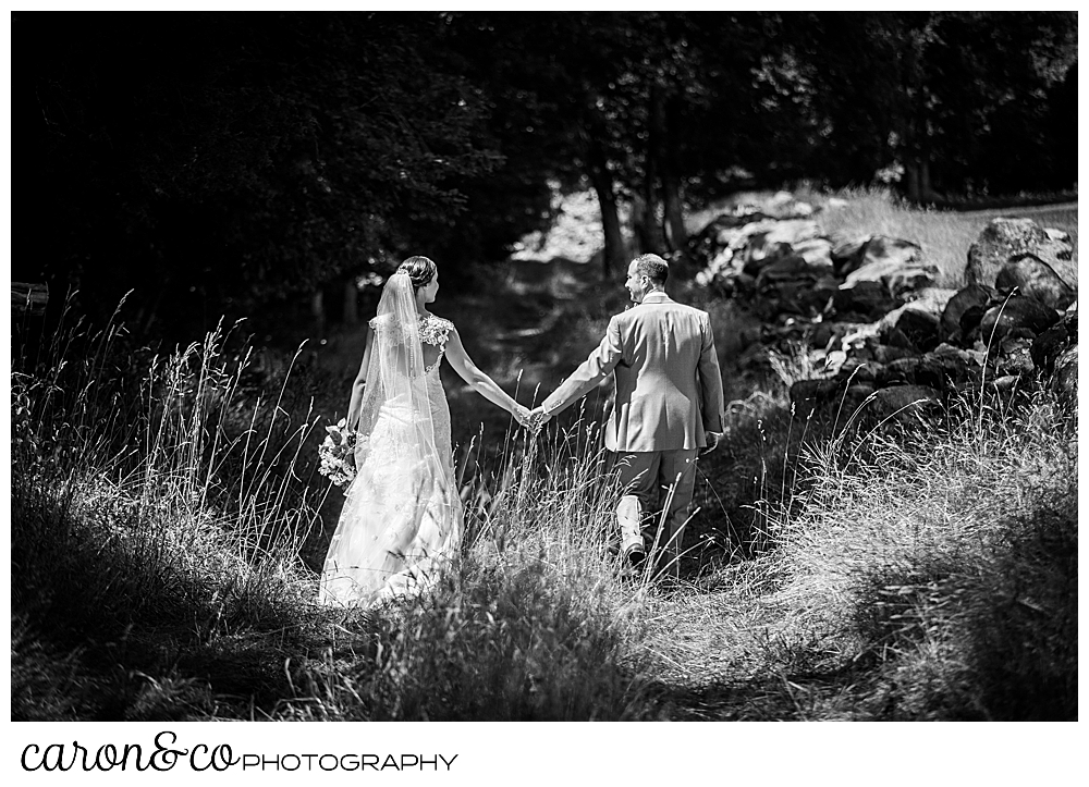 sweet summertime wedding day black and white photo of a bride and groom walking down a grassy lane holding hands