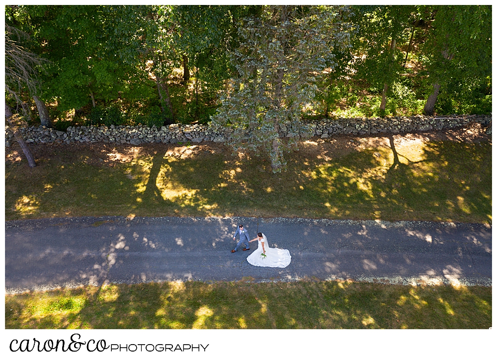 sweet summertime drone wedding photo of a bride and groom walking hand in hand on a shady drive 