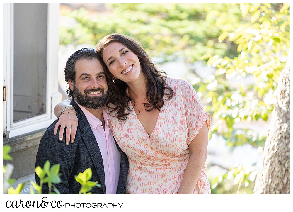 man and woman sitting together, looking toward the camera, the sun is shining in the background of this maine engagement session