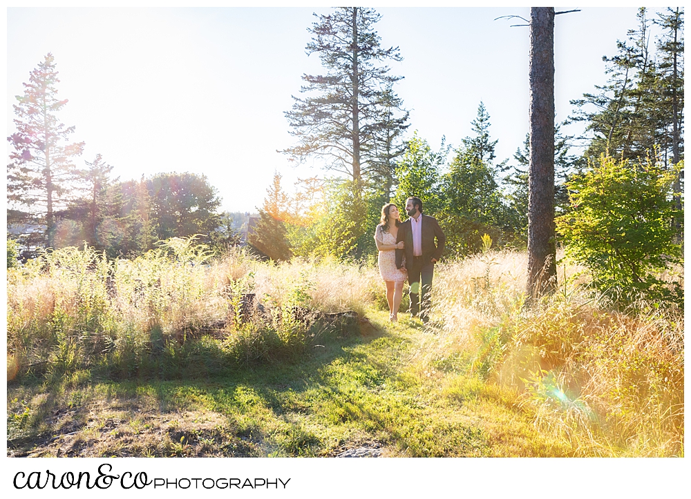 a woman and man walk together in a field with the sun behind them, during a Midcoast Maine engagement