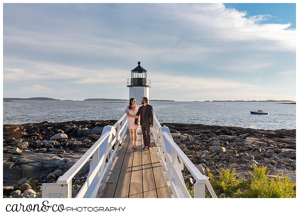 a couple is walking together towards the Marshall Point Lighthouse at their Port Clyde Maine engagement session