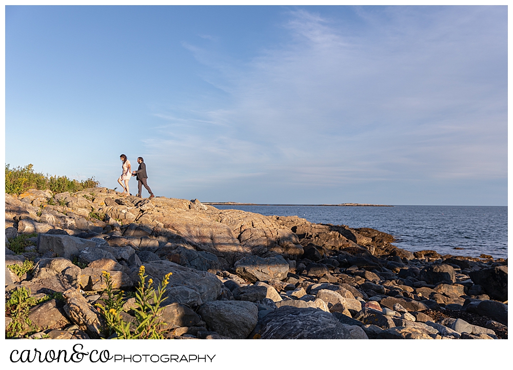 a man and woman are walking on the rocks during their coastal Maine engagement photo session