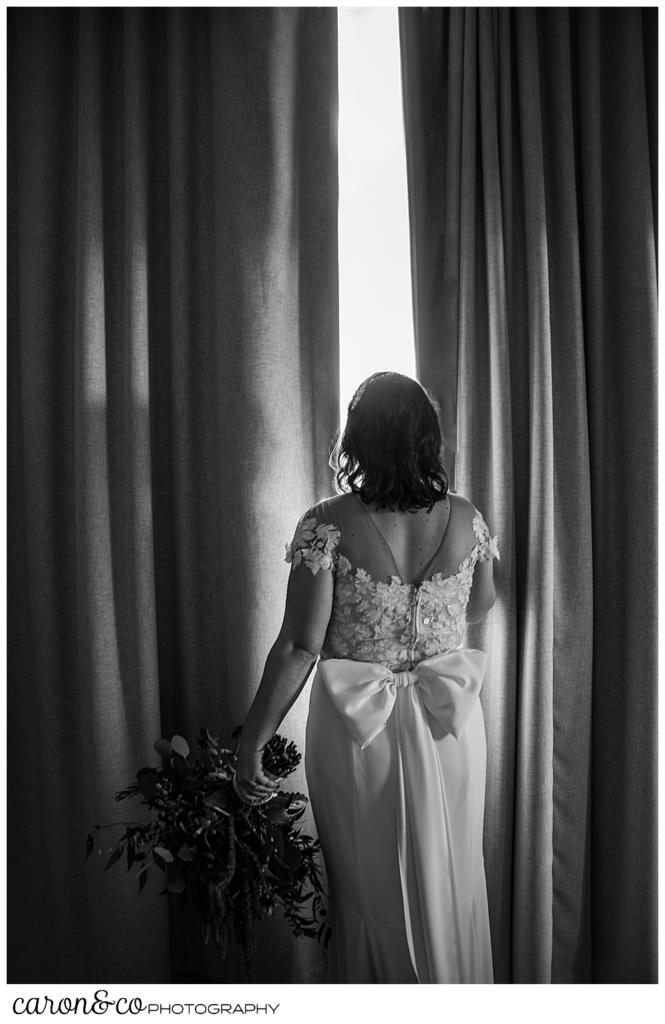 a black and white photo of a bride looking out between two drapes in a hotel room