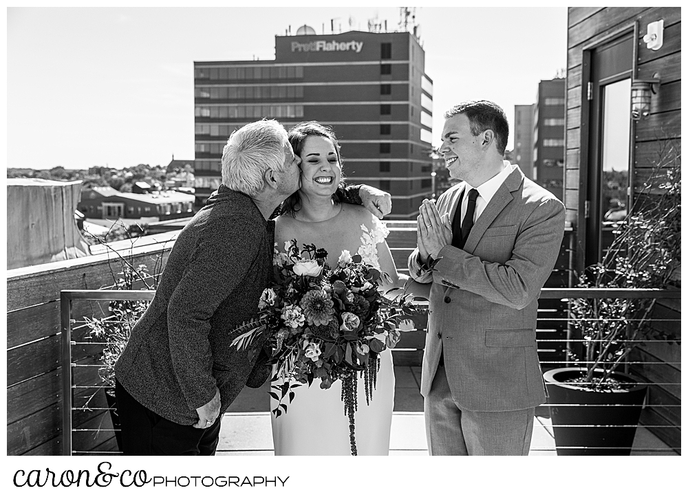 the officiant and relative of the bride, kisses the bride while the groom looks on from the rooftop of their Portland Press Hotel wedding