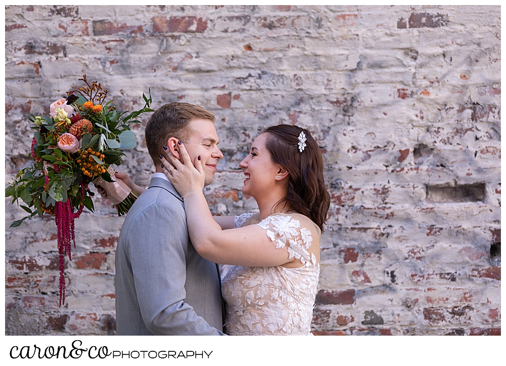 a bride and groom are standing face to face, the bride's hand is on the groom's face