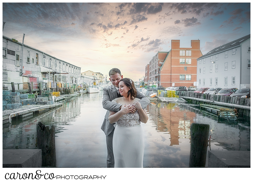 a groom hugs a bride from behind, in front of Portland Maine Harbor