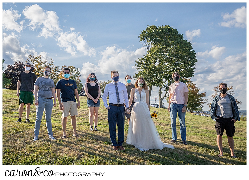 Bride and groom standing on a hill with their friends, they're all wearing masks for cover-19