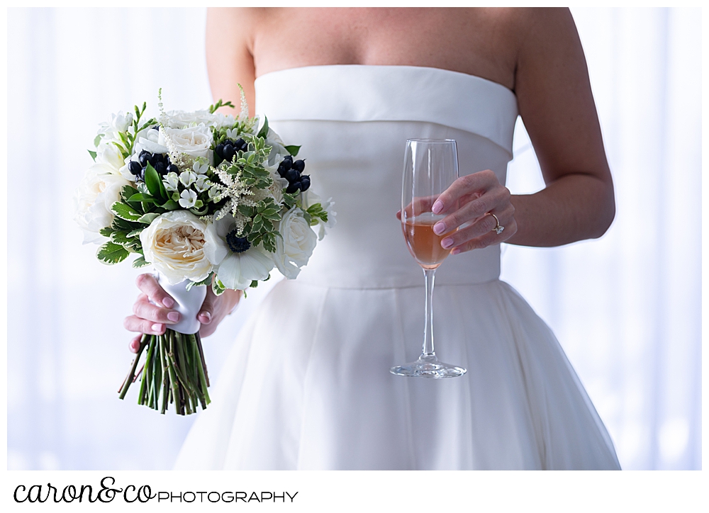 a bride stands holding a beautiful bouquet in one hand, and a glass of champagne in the other, you can't see her head