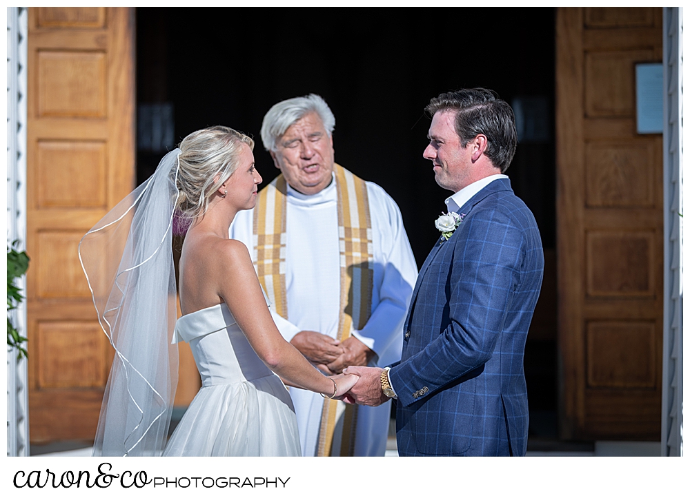 A bride and groom stand before a Catholic priest during their timeless Boothbay Harbor wedding in Maine