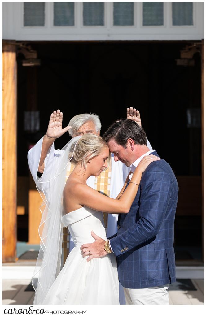 a bride and groom stand with foreheads together, as a priest blesses them at an Our Lady Queen of Peace, timeless Boothbay Harbor wedding