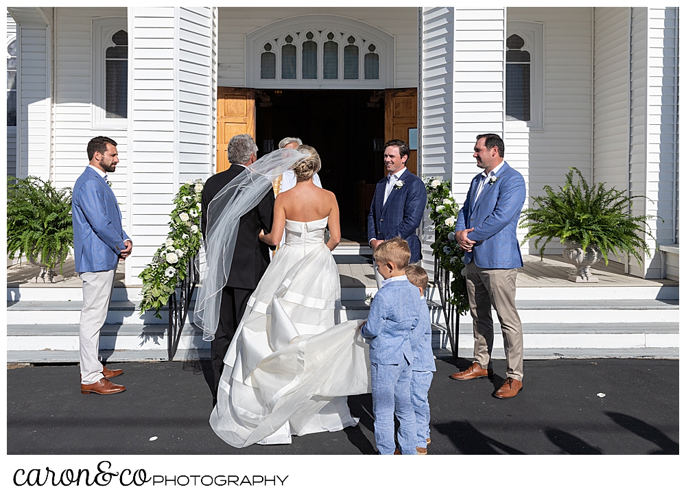 A bride, with two pages carrying her train, and her father approach the front of Our Lady Queen of Peace Catholic Church during a timeless Boothbay Harbor wedding ceremony