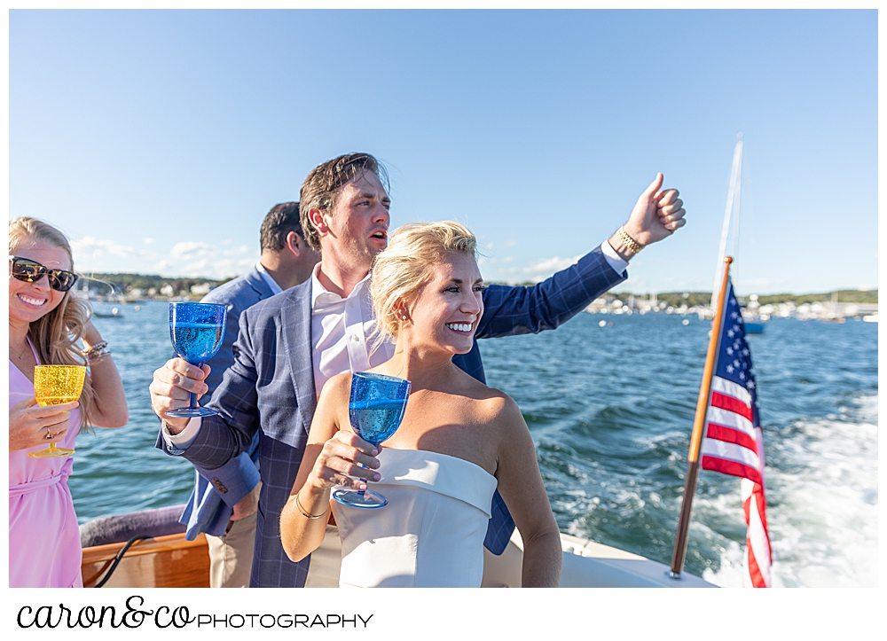 A bride and groom on the back of a Hinckley Yachts Picnic boat, they're holding champagne and waving, during their Boothbay Harbor, Maine wedding