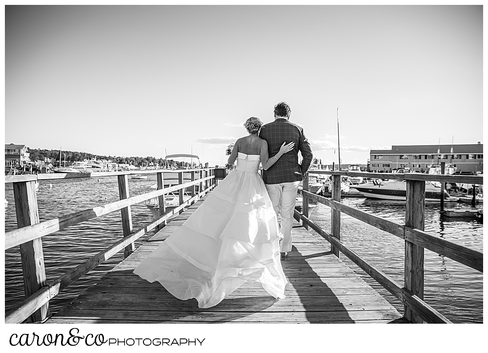 Black and white photo of a bride and groom walking on a dock, their backs are to the camera, the bride's dress is billowing out