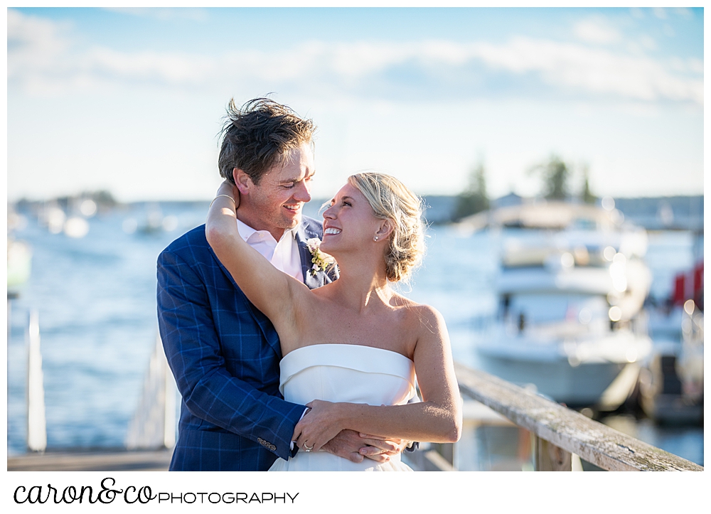 bride and groom standing together, her hand is around his neck, Boothbay Harbor is in the background, during their Boothbay Harbor, Maine wedding