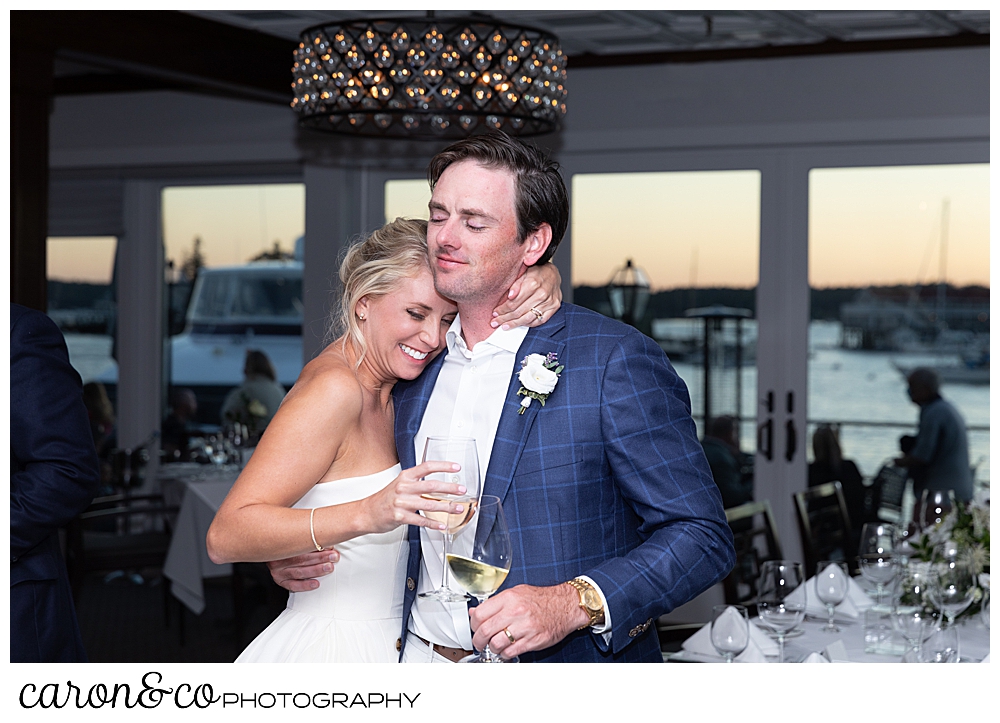 a bride and groom hugging, eyes closed, holding wine glasses during their timeless Boothbay Harbor wedding, Maine