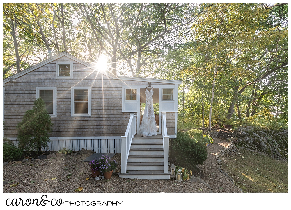 a wedding dress hangs from the doorway of a small cottage, the sun is peeking through the trees behind 