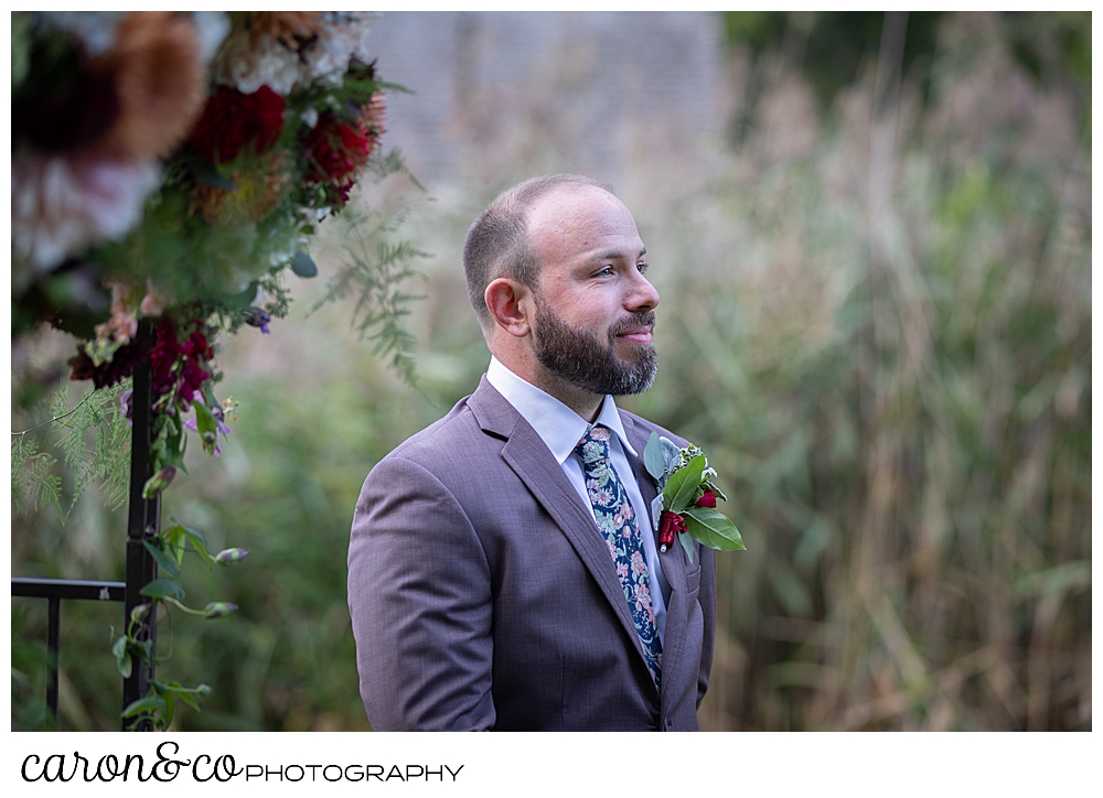 a groom wearing a brown suit, with a flowered tie, stands waiting for his bride to walk down the aisle during a charming Kennebunkport Maine wedding ceremony