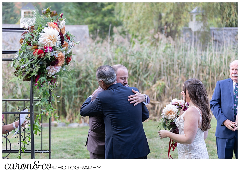 the father of the bride hugs the groom at the flowered arbor at a charming Kennebunkport Maine wedding ceremony