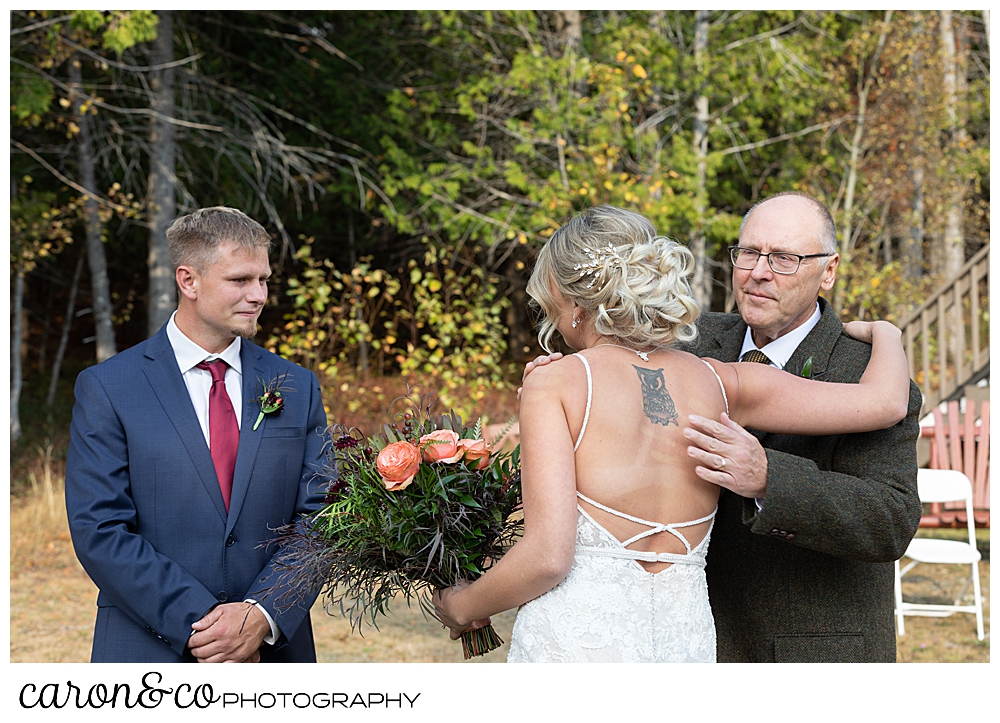 a bride and her father hug, while the groom looks on, at a Rangeley Maine wedding ceremony