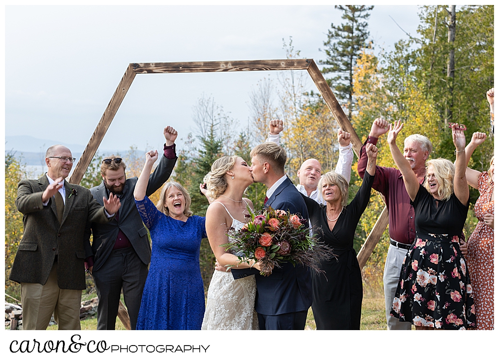 a bride and groom kiss while standing in front of their cheering families during their Rangeley Maine wedding