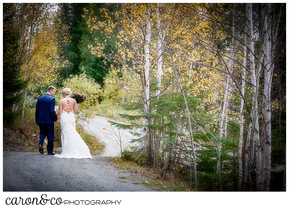 a bride and groom walk, with their backs to the camera, hand in hand down a dirt road in the fall foliage during a Rangeley Maine wedding