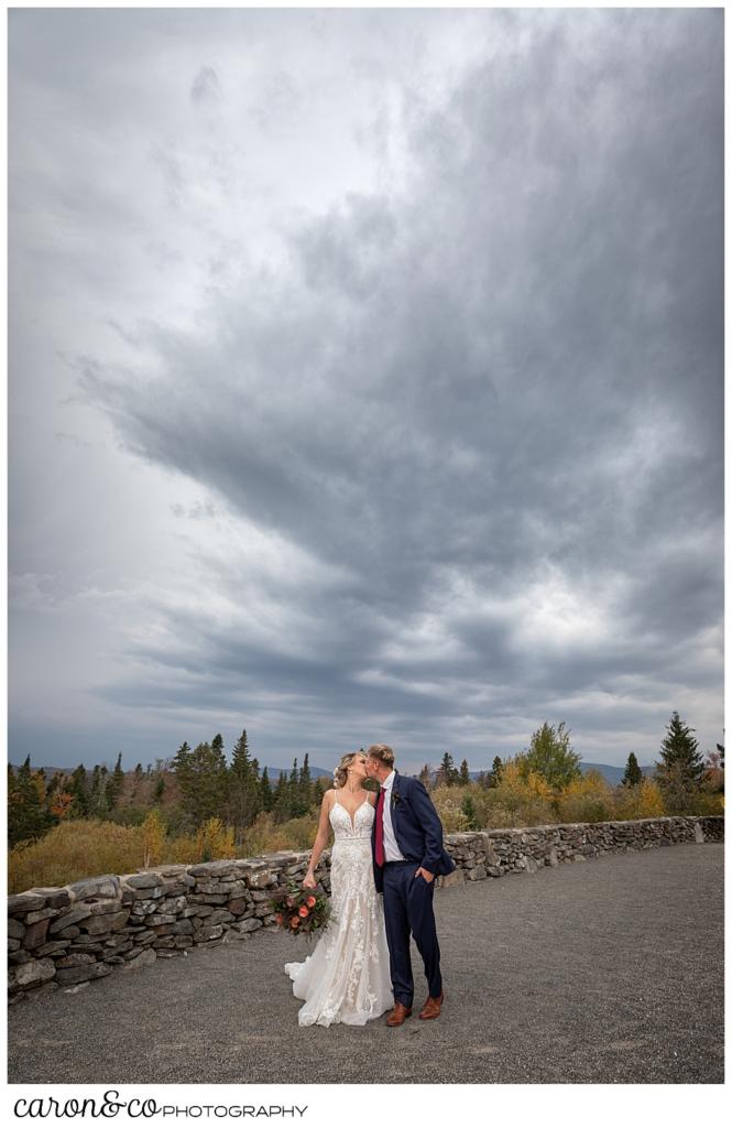 a bride and groom kiss with a dramatic sky behind them at a Rangeley Maine wedding