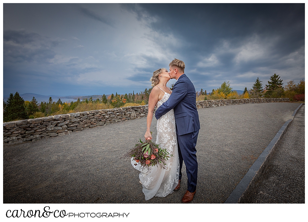 a bride and groom kiss with foliage and dramatic skies in the background during a Rangeley Maine wedding