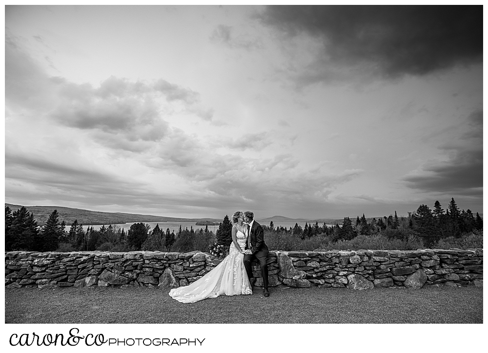 black and white photo of a bride and groom sitting on a stone wall with a dramatic view and skies in the background during their Rangeley Maine wedding