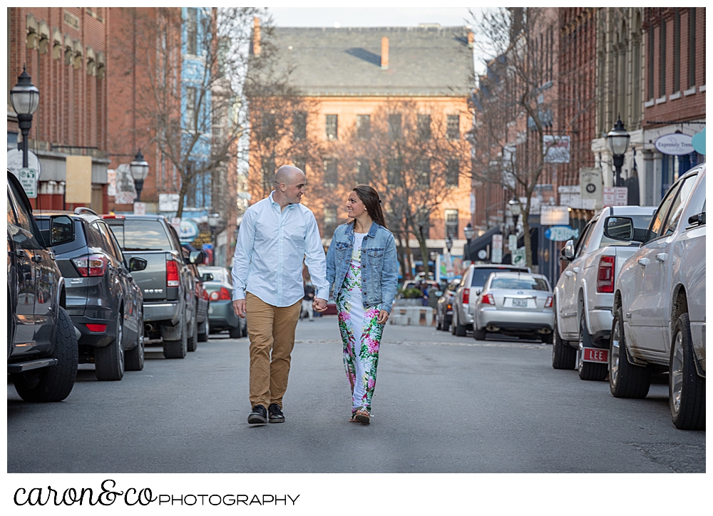 a man and woman holding hands walk up the middle of Exchange Street, Portland, Maine, during their Portland Maine spring engagement photo shoot