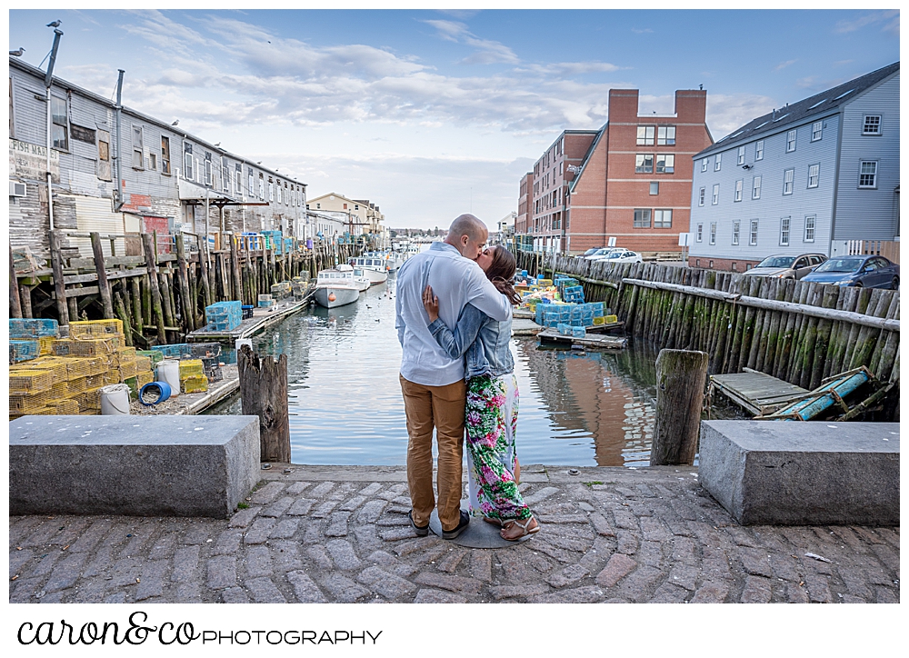 a mand and woman stand with their backs to the camera, kissing, in a walkway between two piers in Portland, Maine, during their Portland Maine spring engagement photo session