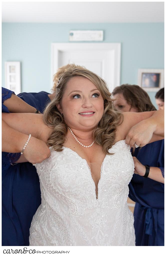 a smiling bride holds up her wedding dress while bridesmaids in blue button the back of her dress