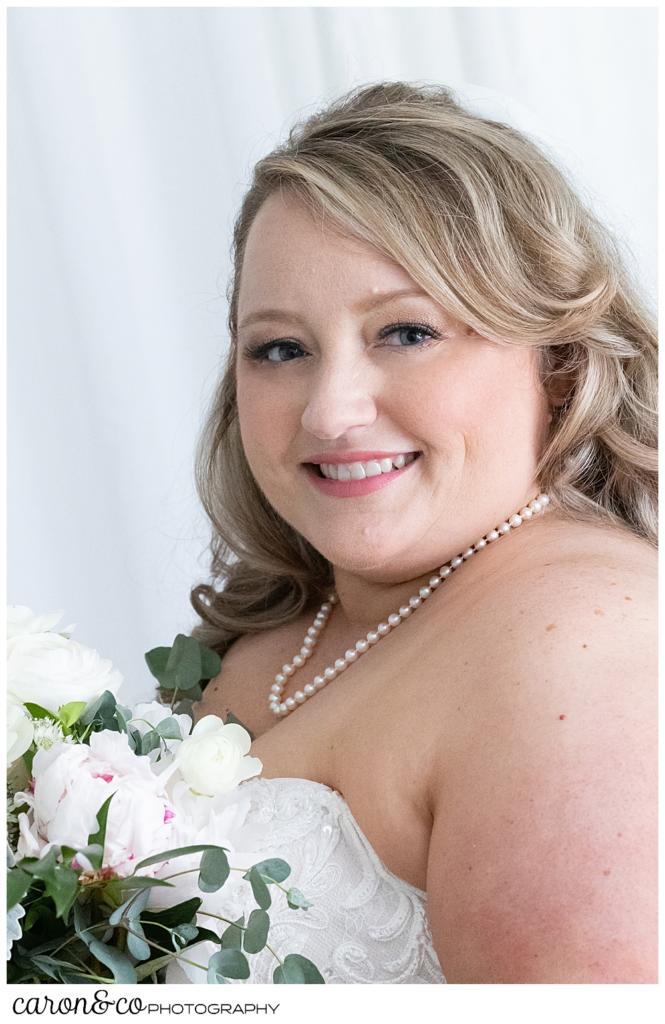 a beautiful bride smiles in her bridal portrait, she is wearing a strapless dress, a pearl necklace, and has white and blush flowers in her bouquet