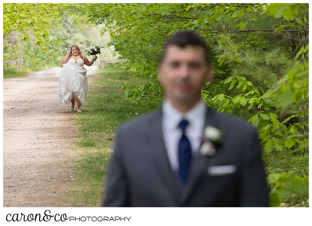a groom stands in the foreground, blurry, as the focus is on the bride, holding up her train, and carrying a bouquet, as she approaches him from behind, on a trail in the woods