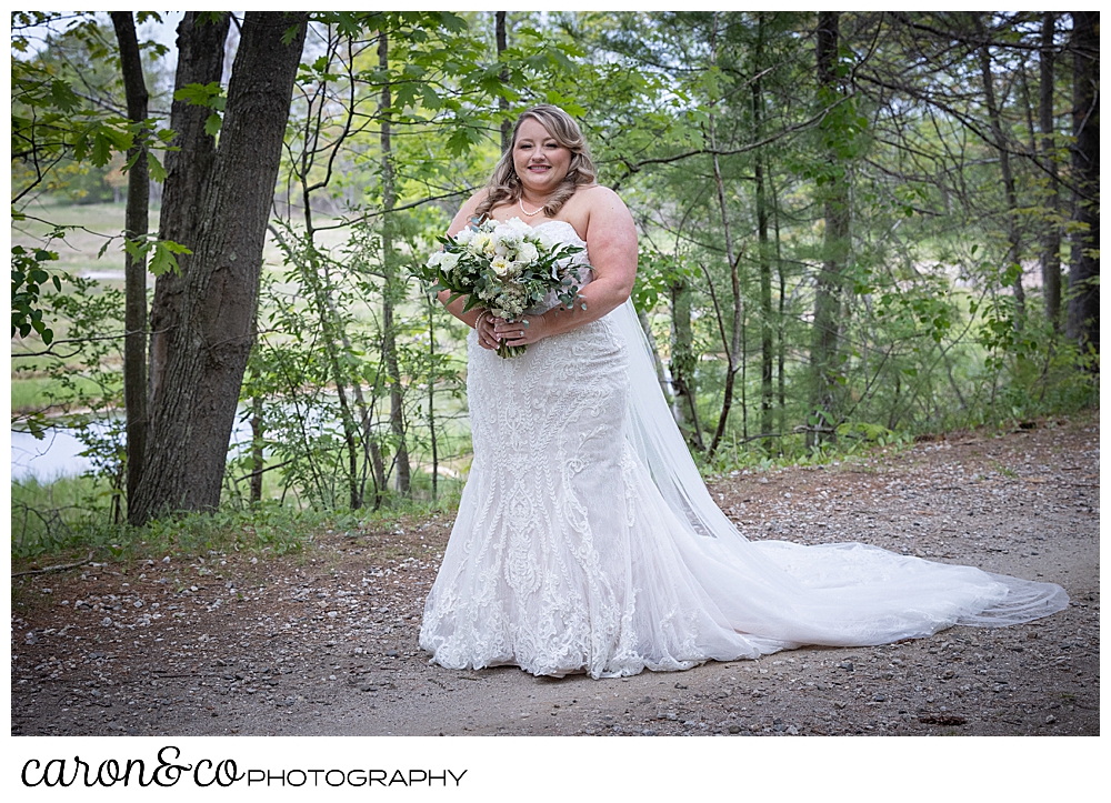 a bride stands on a trail in the woods, she's wearing a strapless white wedding dress and veil, and carrying a bouquet of white, blush and green