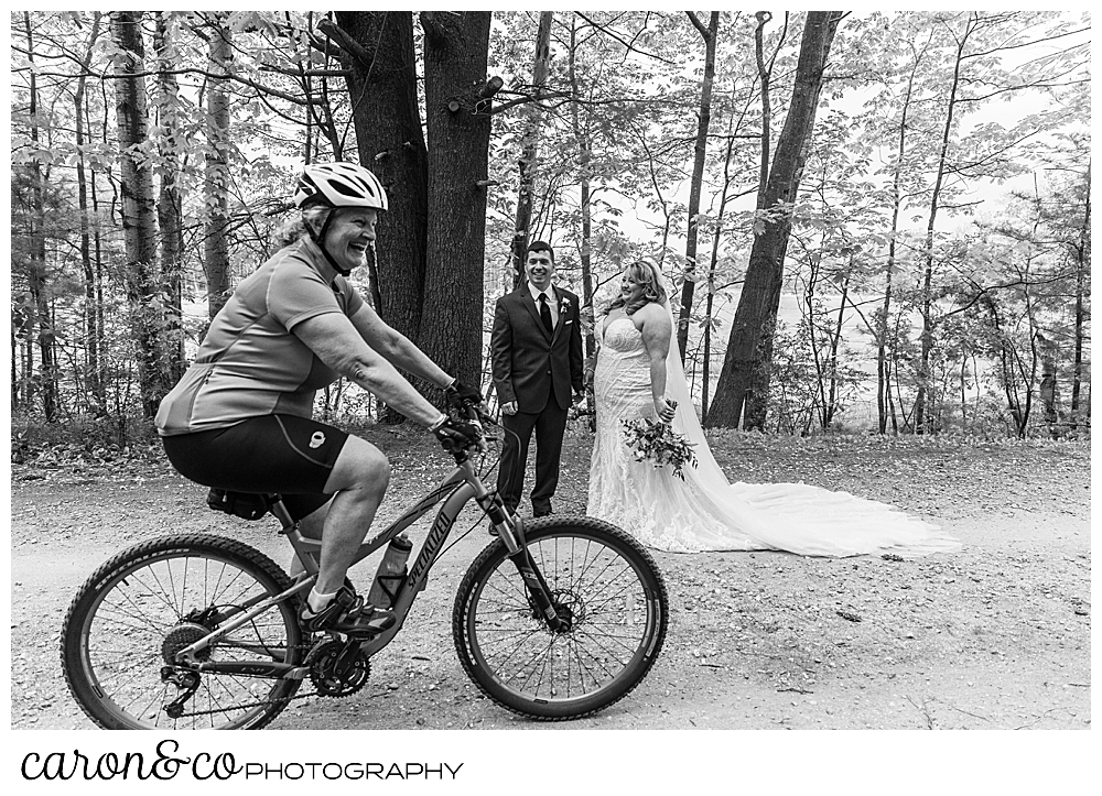 black and white photo of a bride and groom standing together on a path in the woods, as a cyclist goes by in the foreground