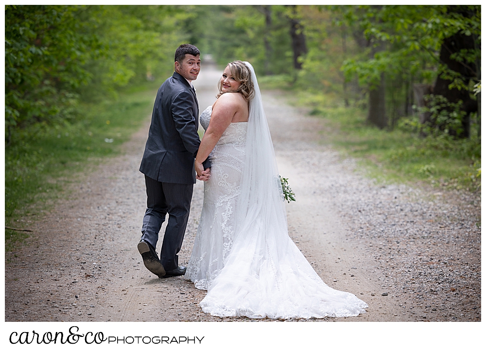 a bride and groom walking on a wooded path, turn to look at the camera