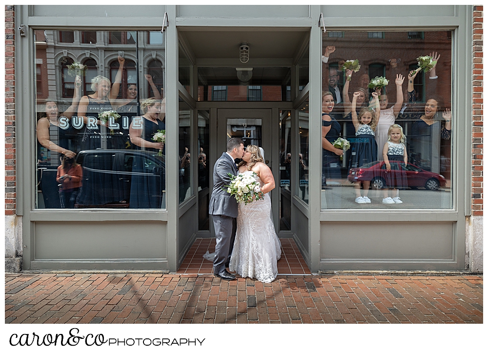 a bride and groom kiss in front of Sur Lie Restaurant in Portland, Maine, while their bridal party cheers from inside