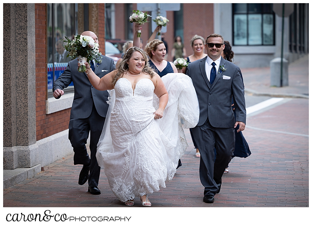 a bride in white, holding a bridal bouquet, walks down the street in Portland, Maine, with her bridal party