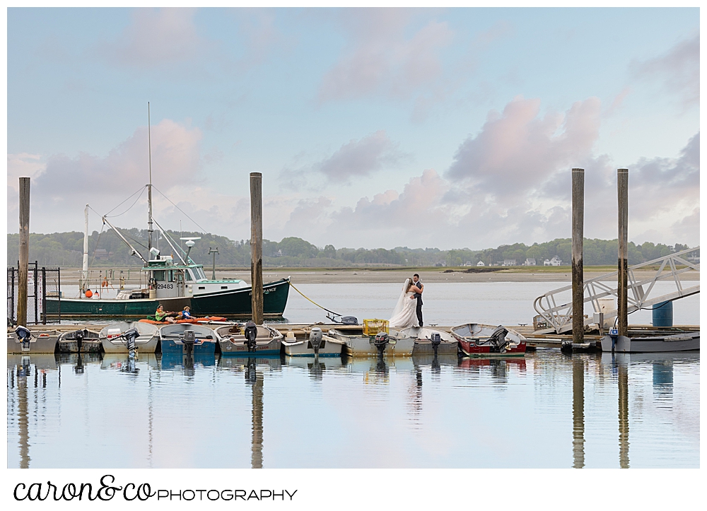 a bride and groom kiss on the docks, with boats and kayaks in the background, at their Pine Point Maine wedding