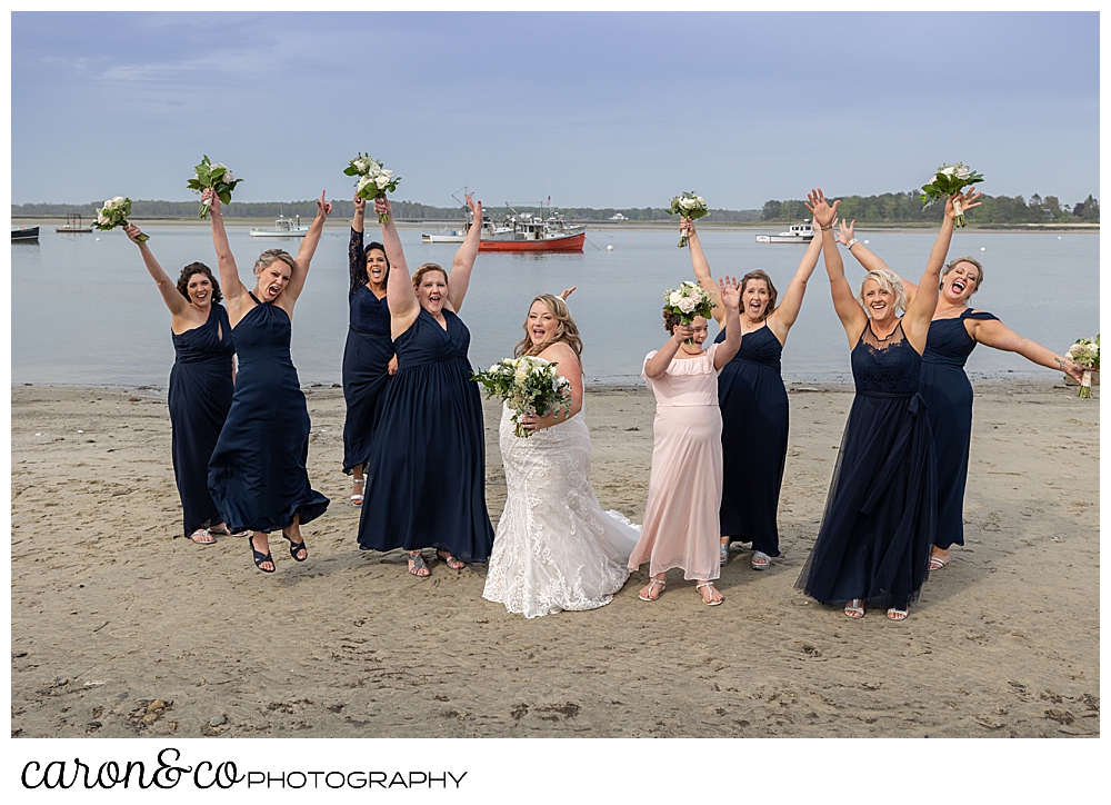 a bride in white, stands with her bridesmaids in blue, as they cheer on the beach at Pine Point Maine wedding