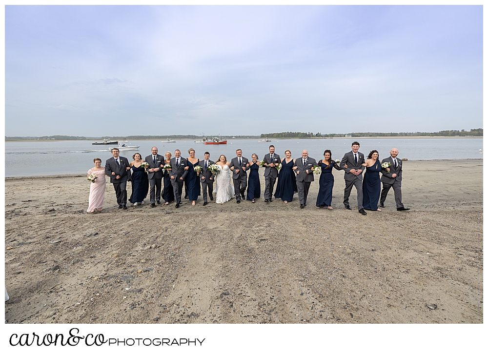 a bride and groom with their bridal party, linked arms and walking on the beach at Pine Point, Scarborough, Maine