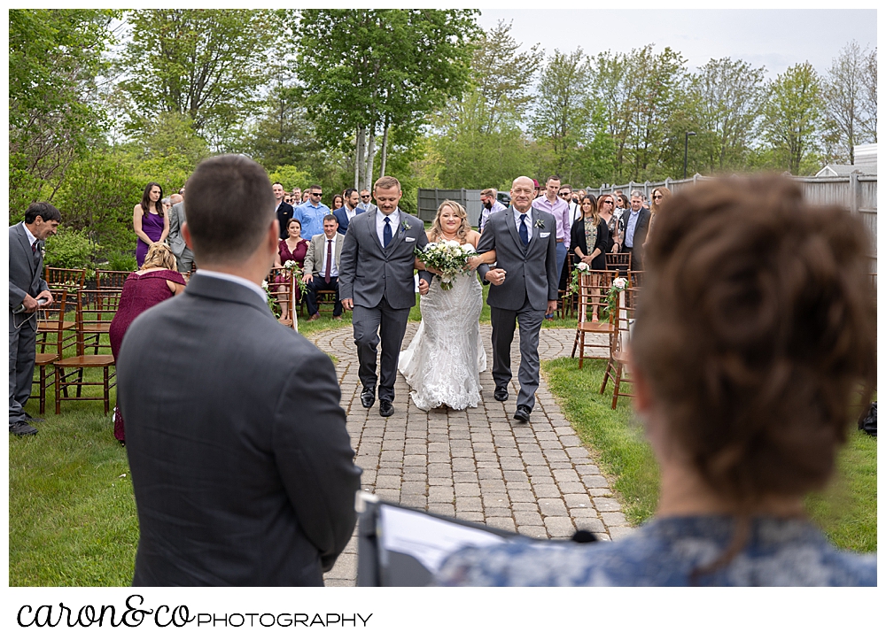 a view from behind the groom and officiant, of a bride being walked down the aisle by her uncle and brother, at a Pine Point Maine wedding