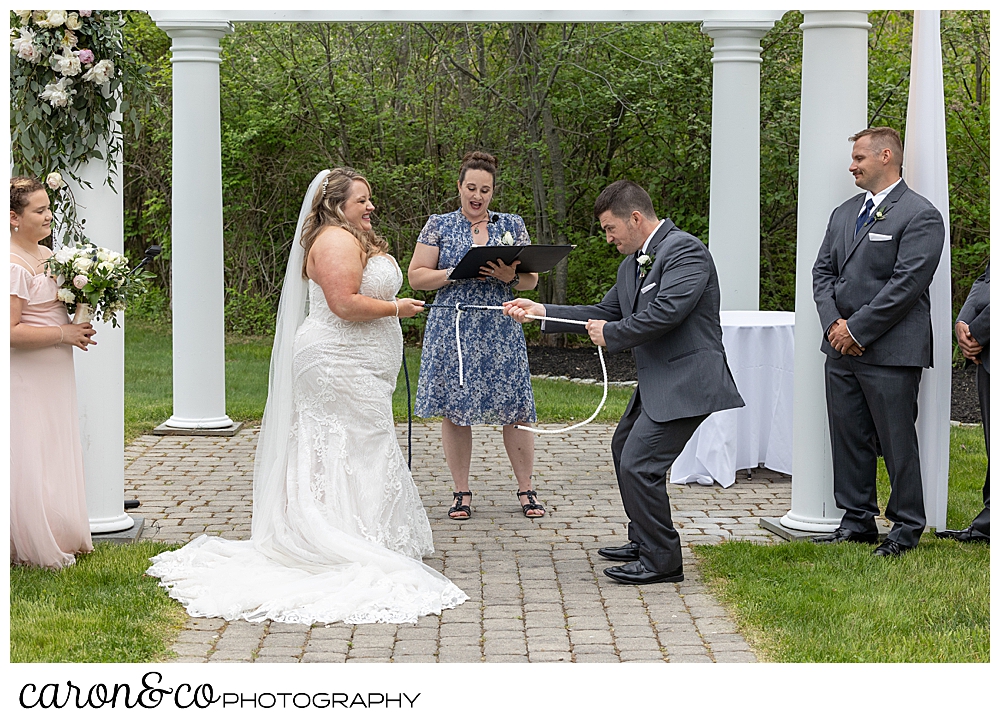 a bride and groom standing before a pergola, and their officiant, complete a knot tying ceremony, as they each pull the secure the ropes at their Pine Point Maine wedding ceremony at the Landing at Pine Point