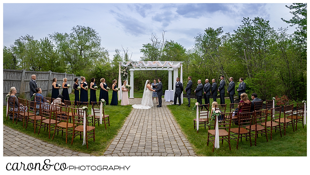 a wide view of a wedding ceremony at the Landing at Pine Point Scarborough, Maine, with the bride, groom, and officiant under the pergola, and the bridal party on either side