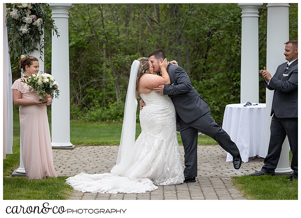 a groom reaches in for the first kiss in front of the pergola at a Landing at Pine Point Maine wedding ceremony