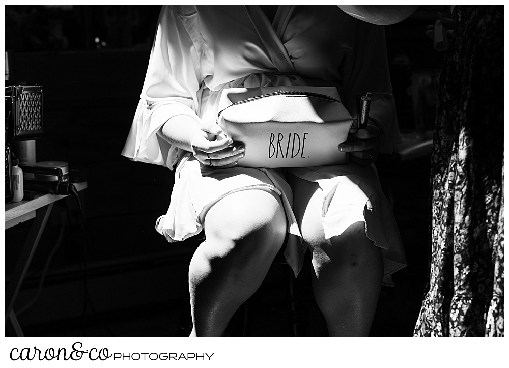 black and white photo of a bride, wearing a robe as she sits in a chair getting her makeup done. She's holding a toiletry bag that says bride