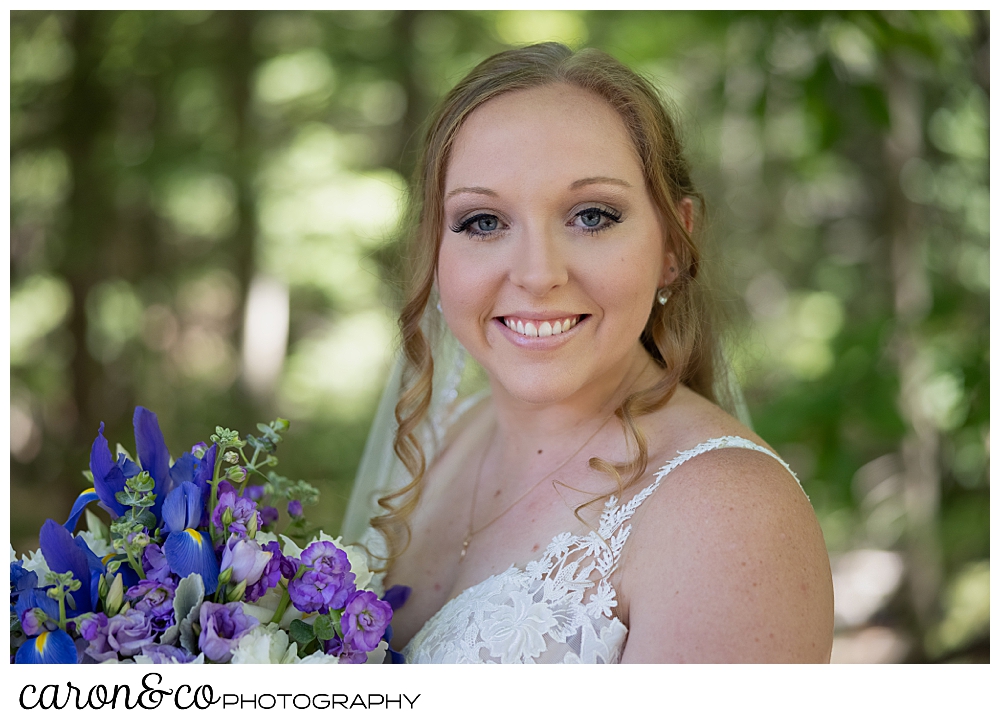 a beautiful bride smiles at the camera, she's wearing a sleeveless dress, and carrying blue and purple flowers